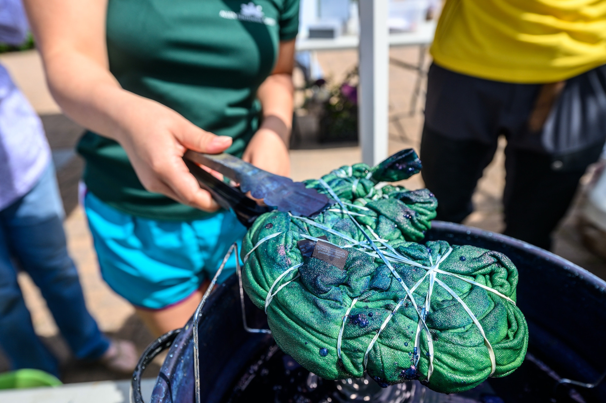 A person holds a tie-dyed tshirt with a pair of tongs after pulling the clothing piece from a large metal pot filled with dark blue liquid.