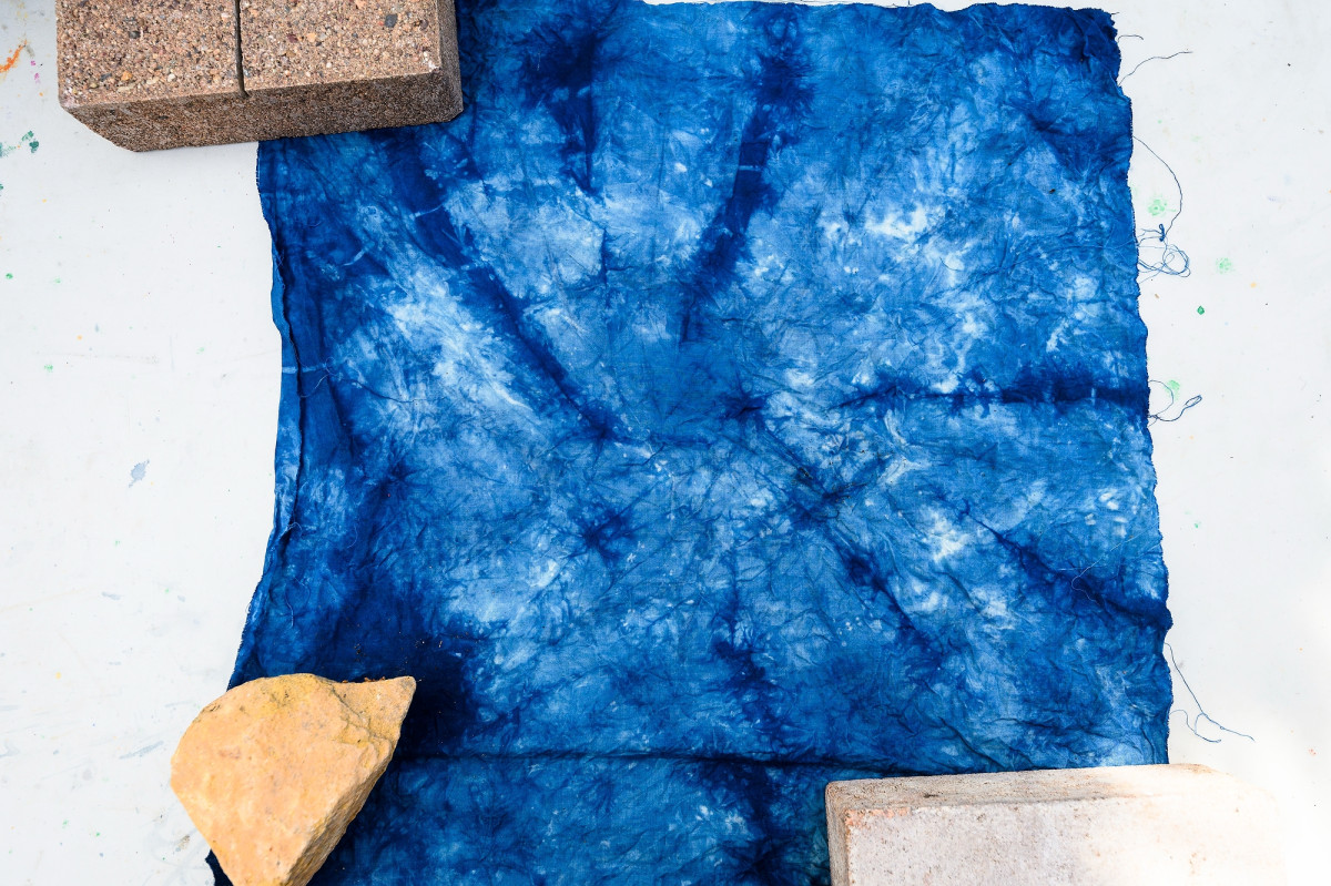 A piece of blue tie-dyed fabric is held by bricks and rocks as it lays out to dry on a table.
