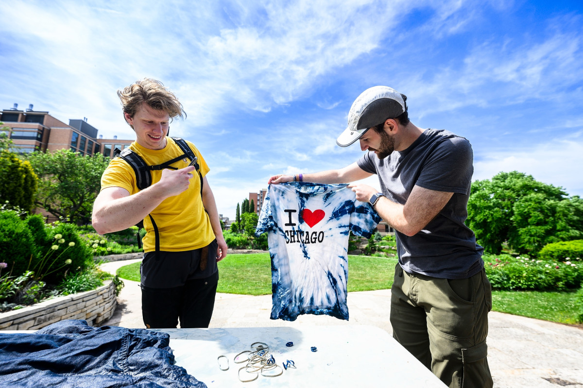 A person holds up an unfolded piece of tied-dyed clothing while another person takes a picture of the finished piece with their phone.