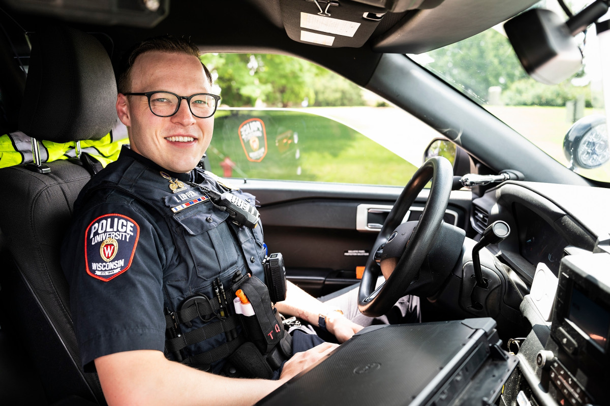 Jake Lepper sits behind the wheel of a police car wearing a UW Police Department uniform.