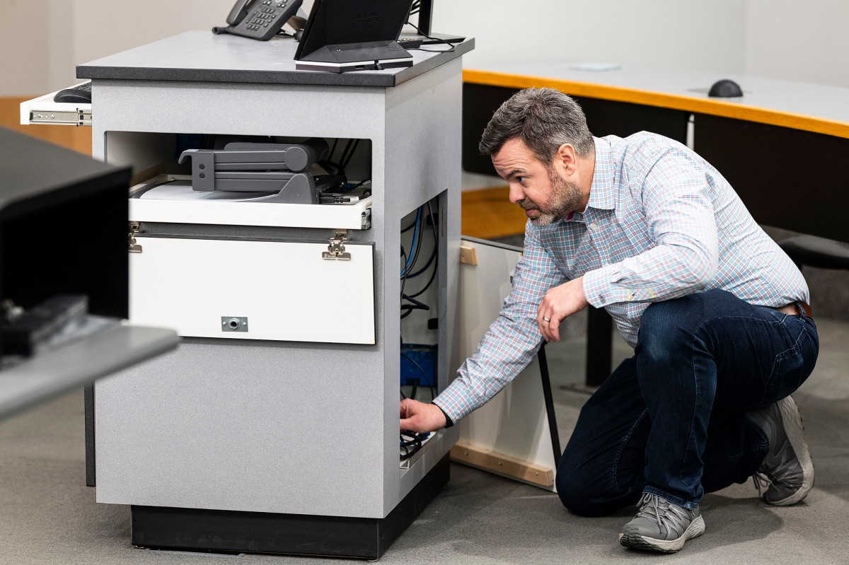 Patrick Long kneels on the floor of a classroom as he looks into a cabinet holding audio-visual equipment.