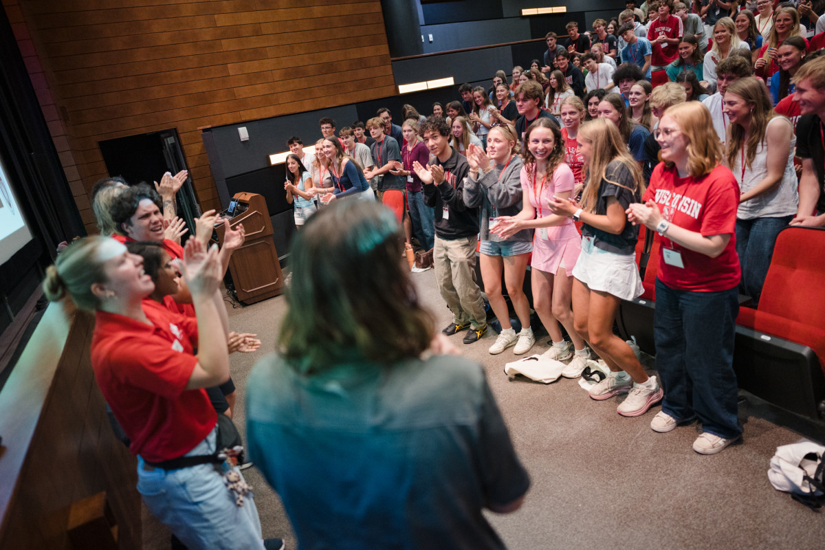 A row of UW–Madison students, all wearing bright Badger red shirts, stand in front of a full auditorium and lead the room of new students in a clapping chant. 