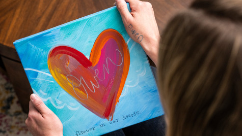 Person holding a colorful painted canvas featuring a large heart design with the word 'Owen' and the phrase 'forever in our hearts'.