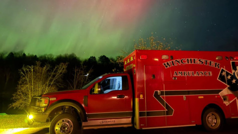 A red ambulance is parked on a rural road at night as Northern Lights brighten the sky overhead.