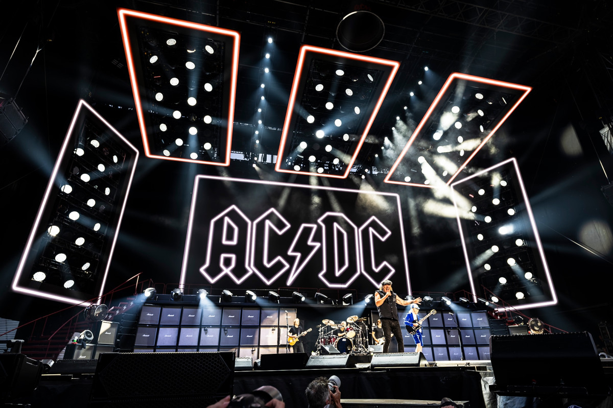 The band ACDC performs on stage in Camp Randall in front of a large neon sign that shows the band's logo.