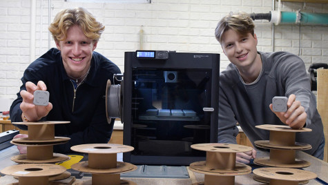 Two young men smile while holding small, square grey devices, standing behind a table with stacks of empty filament spools and a black 3D printer.