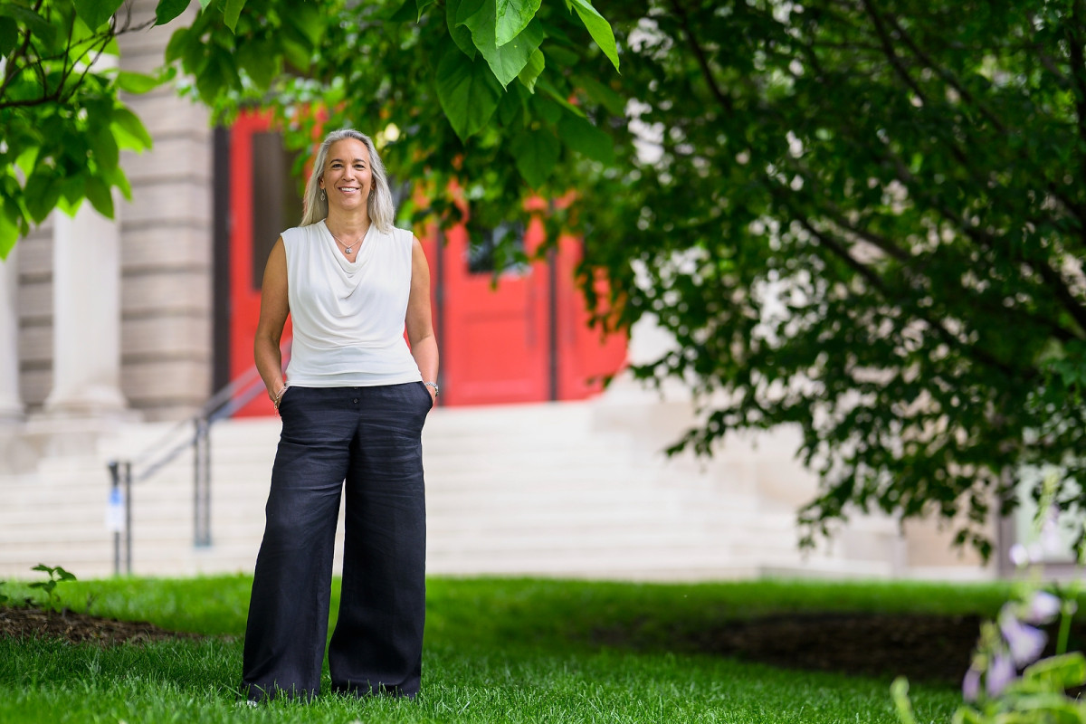 A smiling person standing with hands in their pockets in a grassy area in front of a building with a red door.