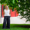 A smiling person standing with hands in their pockets in a grassy area in front of a building with a red door.