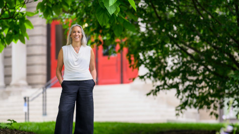 A smiling person standing with hands in their pockets in a grassy area in front of a building with a red door.