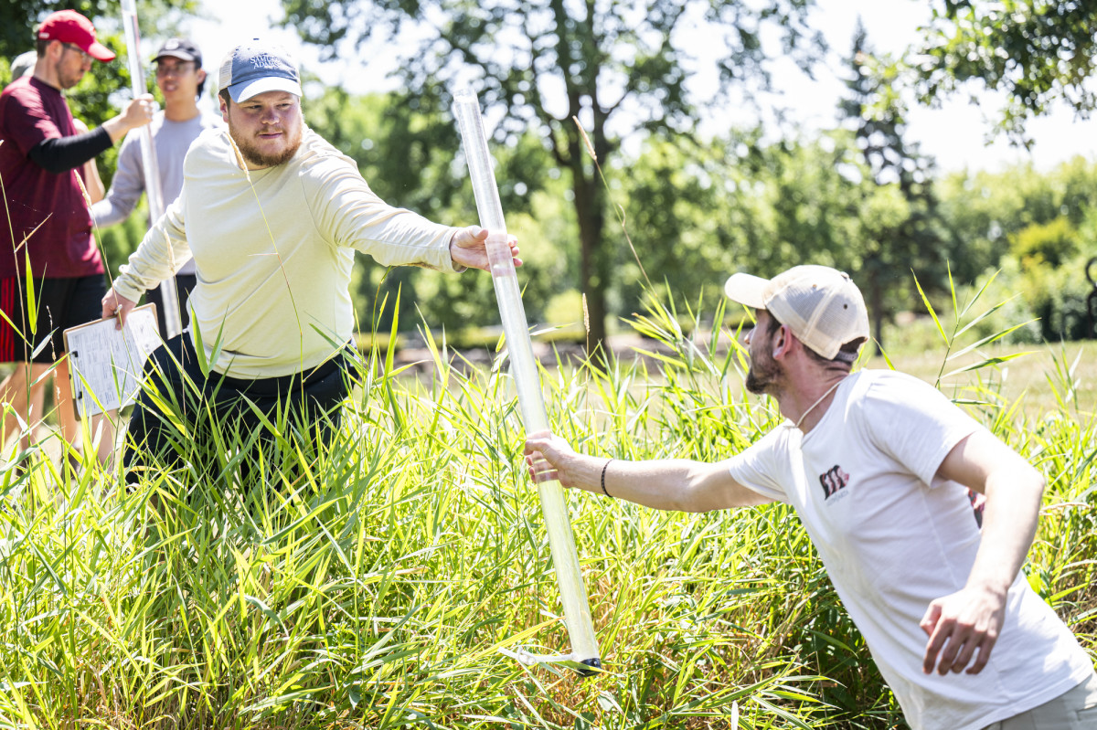 One students stands in the water and hands a sample tube filled with water to another student who stands on the bank of the creek.