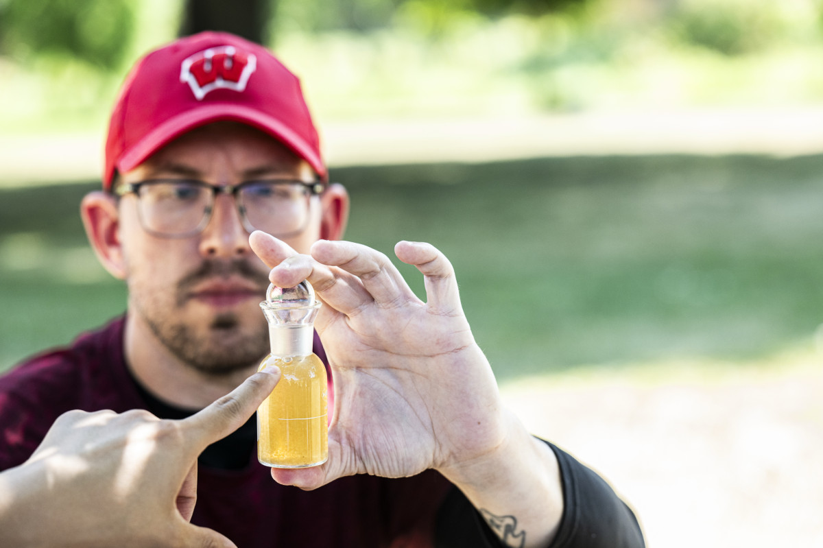 A man wearig a red Wisconsin Badgers hat holds up a glass vial of creek water.