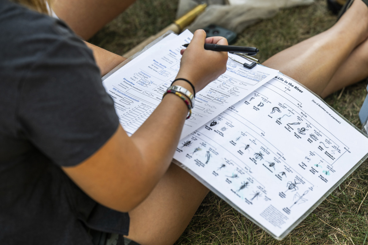 A student sits on the ground and completes paper work detailing what was seen in their water sample.