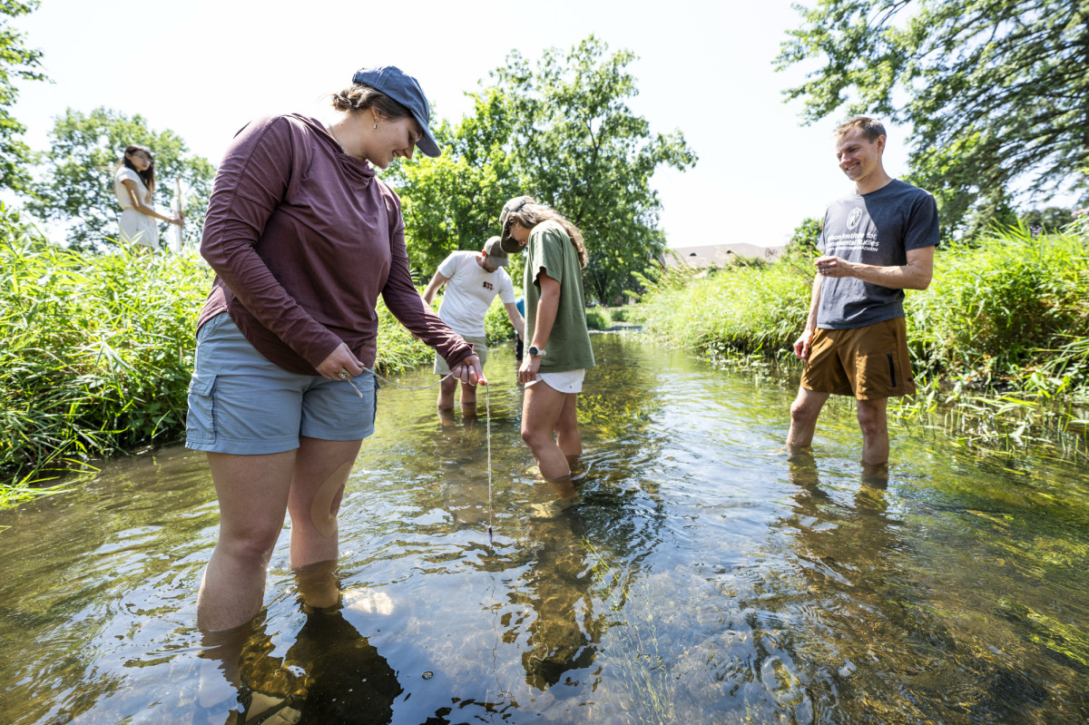 Students stand in a shallow creek. They took down at the water where they are taking samples from.