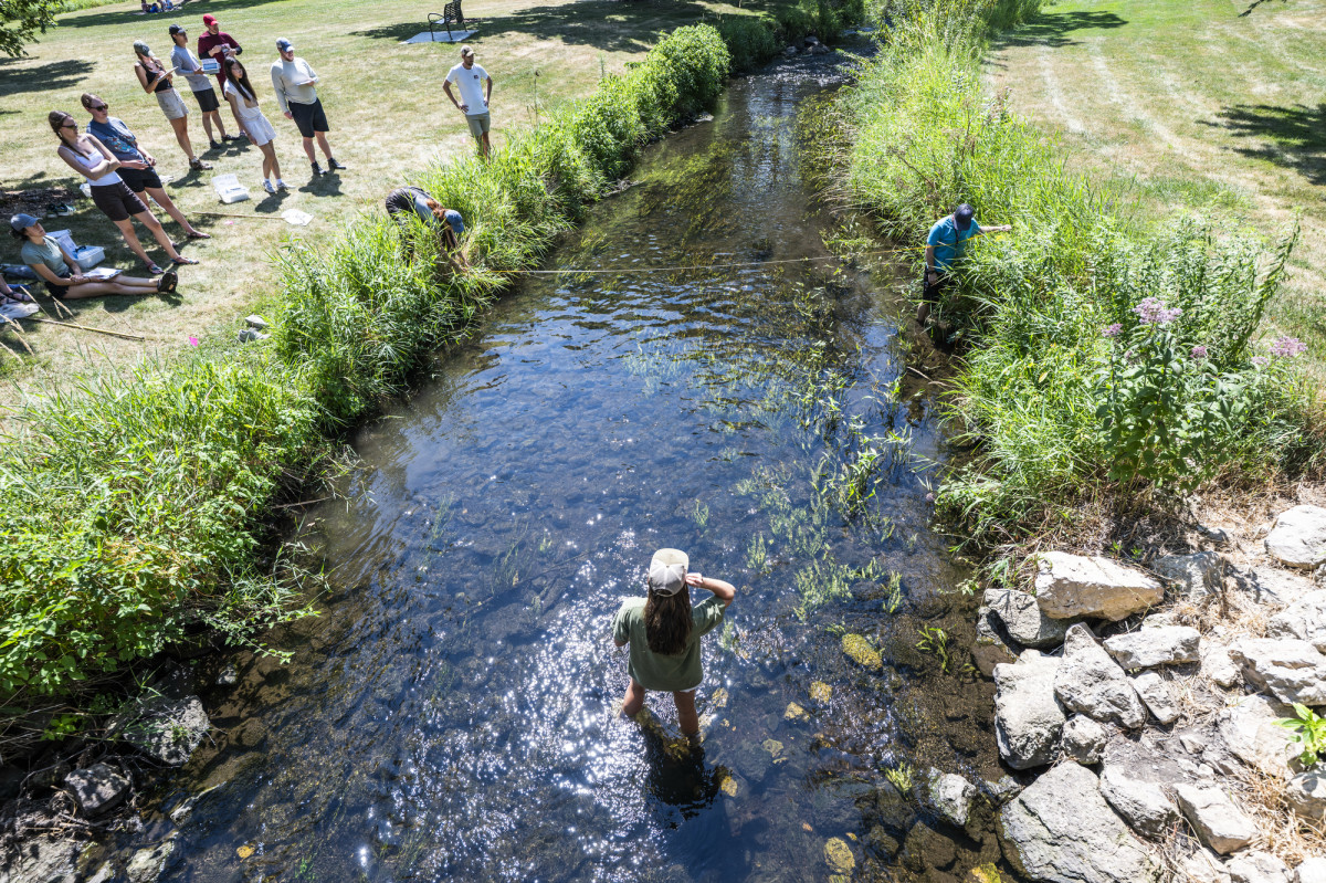 Three students stand in the creek bed, measure the water's width while the other students stand on the grassy land and watch.