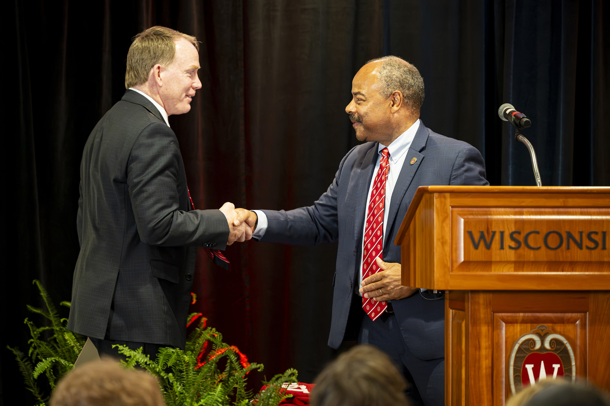 Shawn Eichorst stands on the left and shakes hands with interim Chancellor Eric Wilcots, who stands on the right behind a wooden University of Wisconsin podium. 