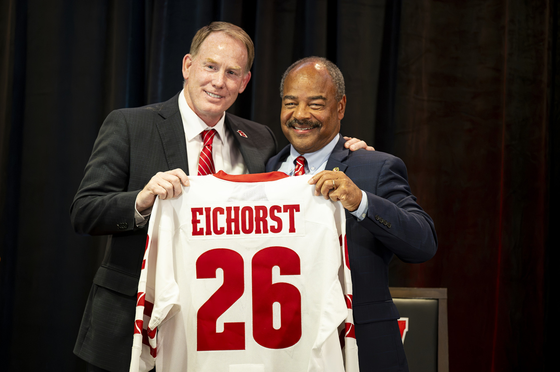Shawn Eichorst on the left and Eric Wilcots on the right stand smiling side-by-side, jointly holding up a white University of Wisconsin women's hockey jersey. The back of the jersey displays the name "EICHORST" in bold red letters above the large red number "26". 