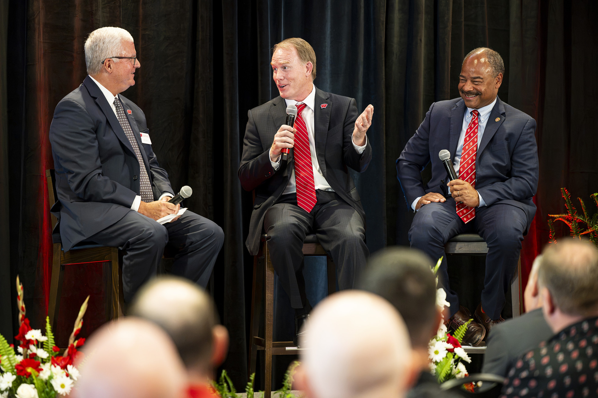 Matt Lepay, Shawn Eichorst and Eric Wilcots sit on high stools on a stage during a panel or Q&A session against a black curtain background. 