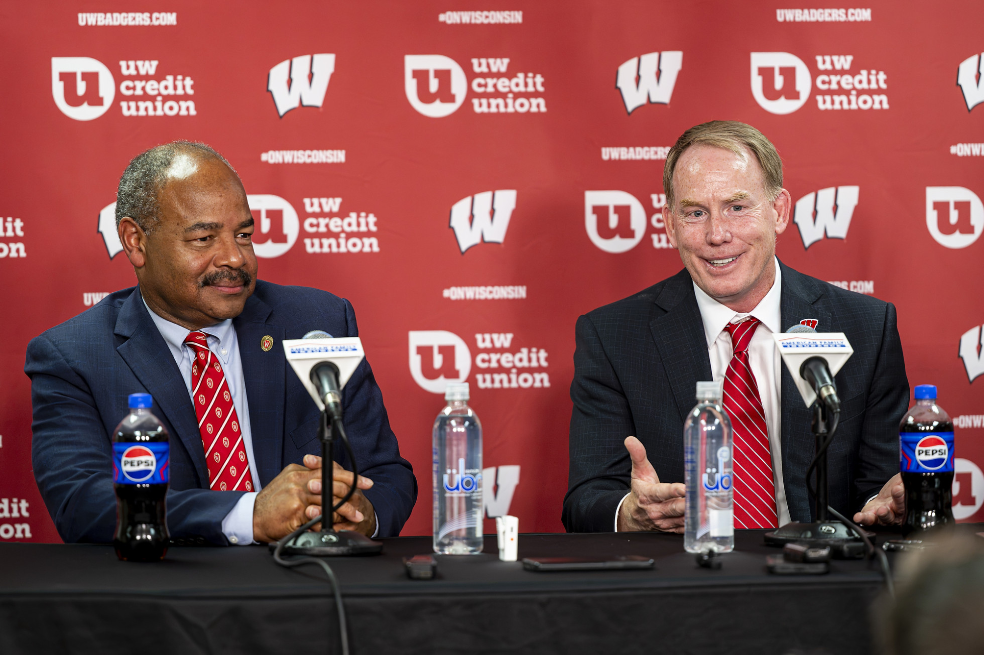 Eric Wilcots and Shawn Eichorst sit at a press conference table, in front of a red backdrop patterned with the Motion W and UW Credit Union branding.
