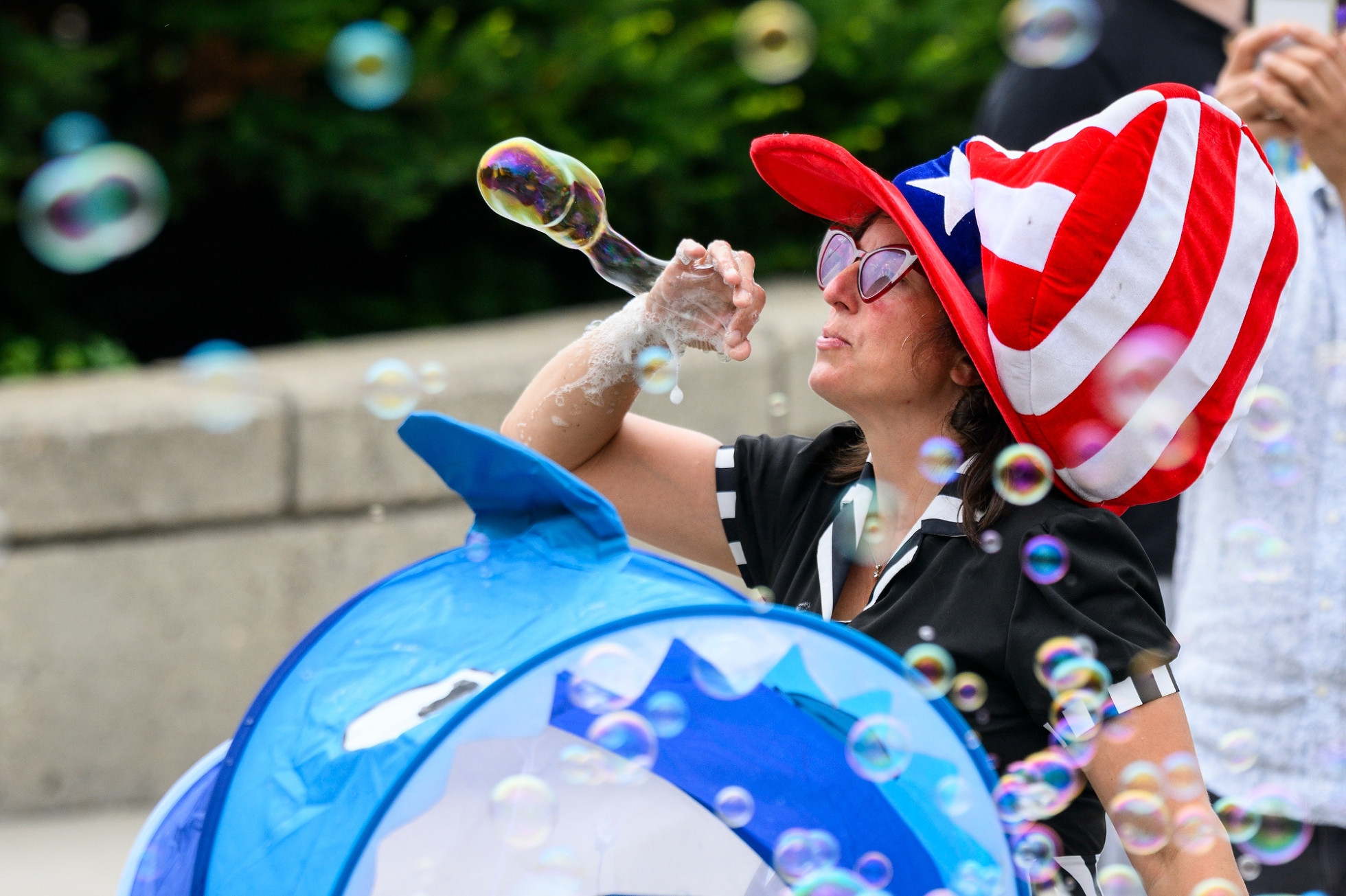 A woman wearing an oversized red, white and blue hat and sunglasses blows a large soap bubble.