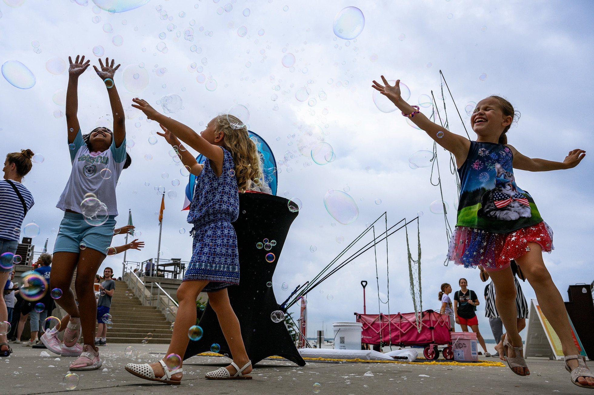 Three children joyfully leap and reach for bubbles on an outdoor concrete plaza.