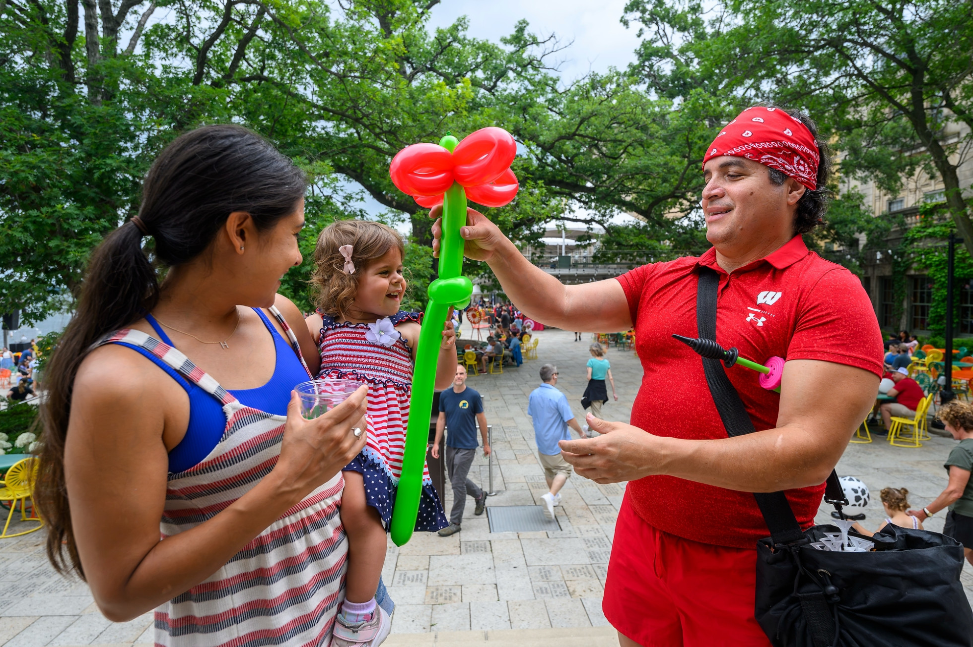 A man wearing a red bandana and a red shirt hands a green and red balloon flower to a smiling toddler on an outdoor plaza. 