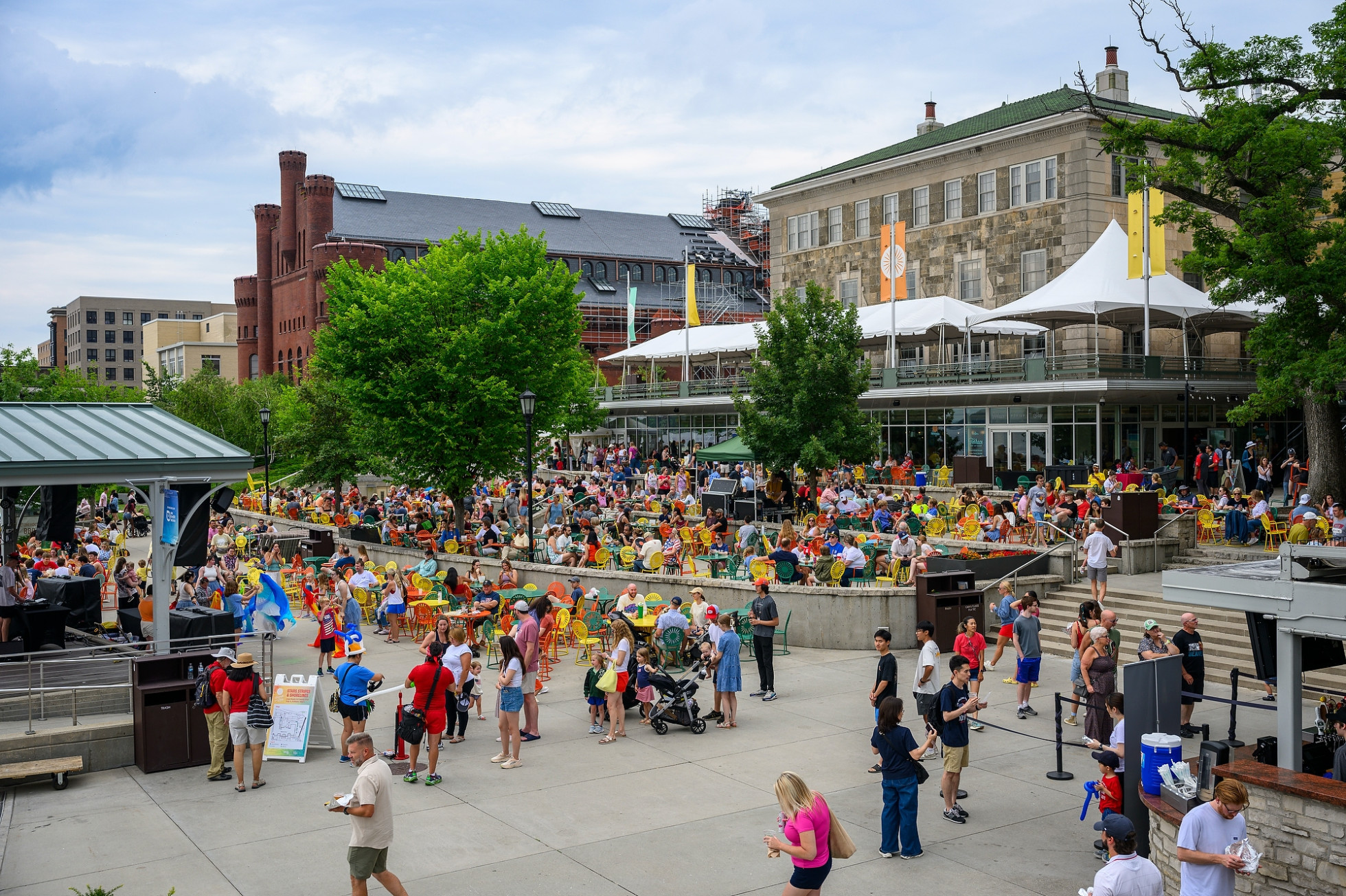 An outdoor terrace plaza filled with people enjoying a summer day. In the midground, numerous people sit at vibrant yellow, orange, and green sunburst-patterned metal tables and chairs, while others walk across the wide concrete foreground. 