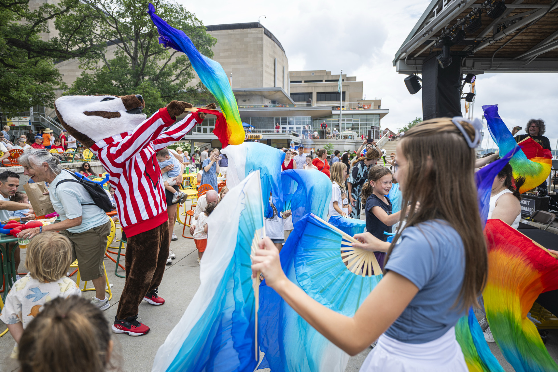A vibrant outdoor event featuring Bucky Badger, surrounded by a lively crowd. Festive, colorful banners and fans in shades of blue and rainbow hues are being waved by attendees. 