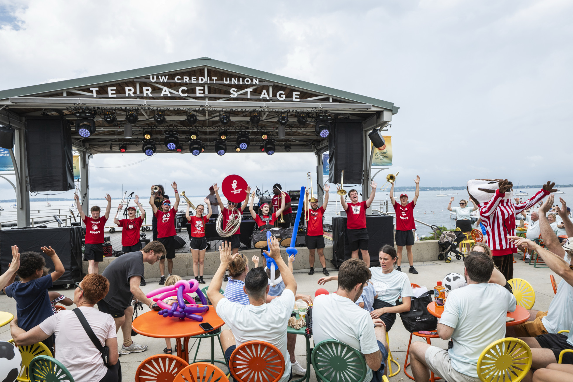 A lively outdoor performance by a marching band on the UW Credit Union Terrace Stage. The marching band in bright shirts are playing instruments and raising their arms, with Bucky joining in. The audience, seated on colorful chairs, watches and claps. Balloons are visible on tables, and boats are in the background on the water.