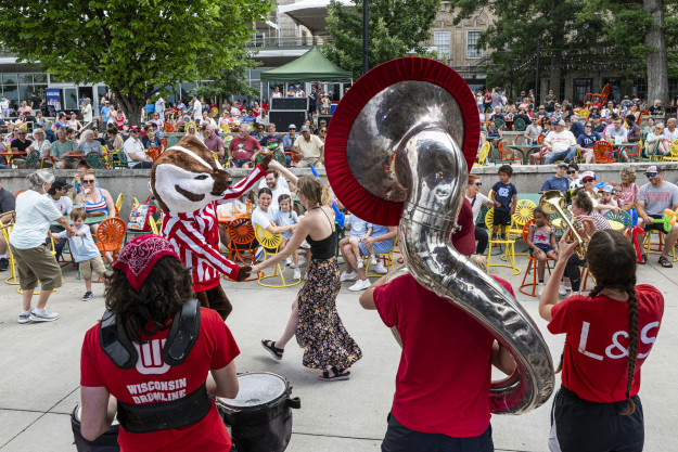 A lively outdoor performance features a marching band with several people playing instruments, including a tuba. Bucky Badger is dancing with an audience member. A seated crowd watches the event in the background, with trees and a building visible.