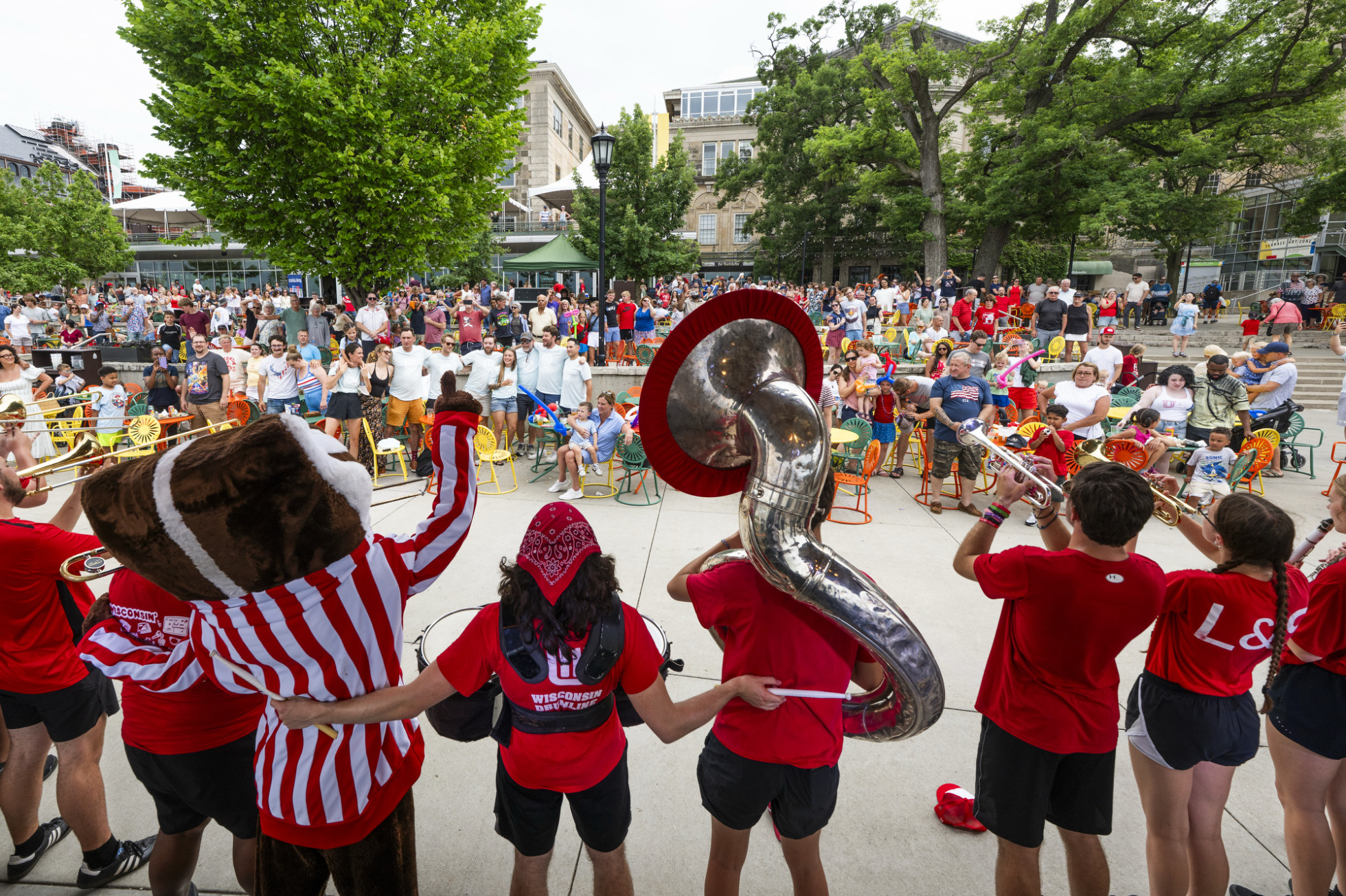 A marching band with Bucky performs and links arms outdoors. A crowd of people watches the performance, seated and standing at the Terrace with trees in the background.
