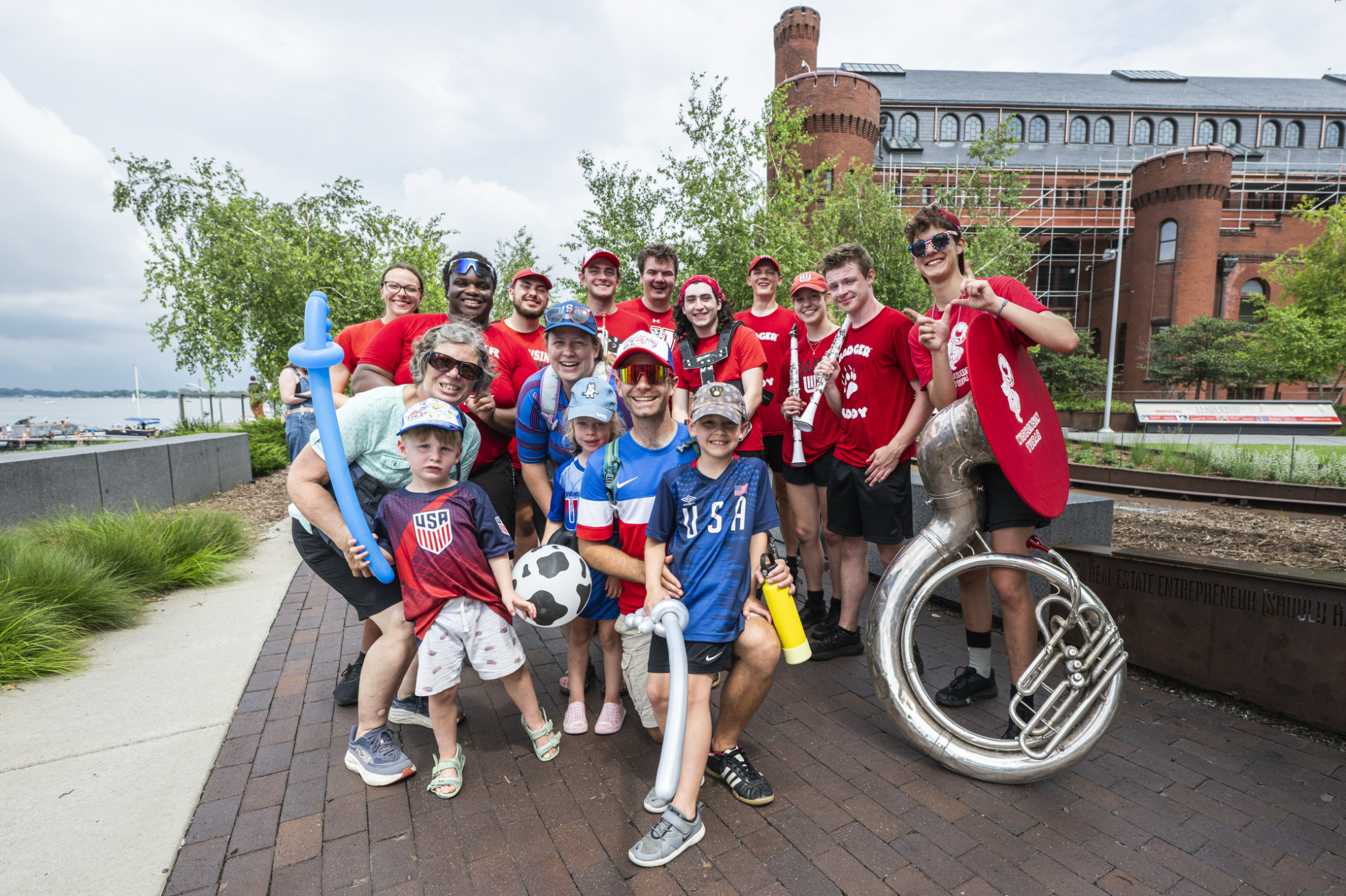 A group of people, including children, pose cheerfully outdoors near the Red Gym in front of Alumni Park. Some are wearing red, white and blue USA soccer apparel. A large silver tuba is held by one individual, and others hold soccer-themed items and balloon swords.