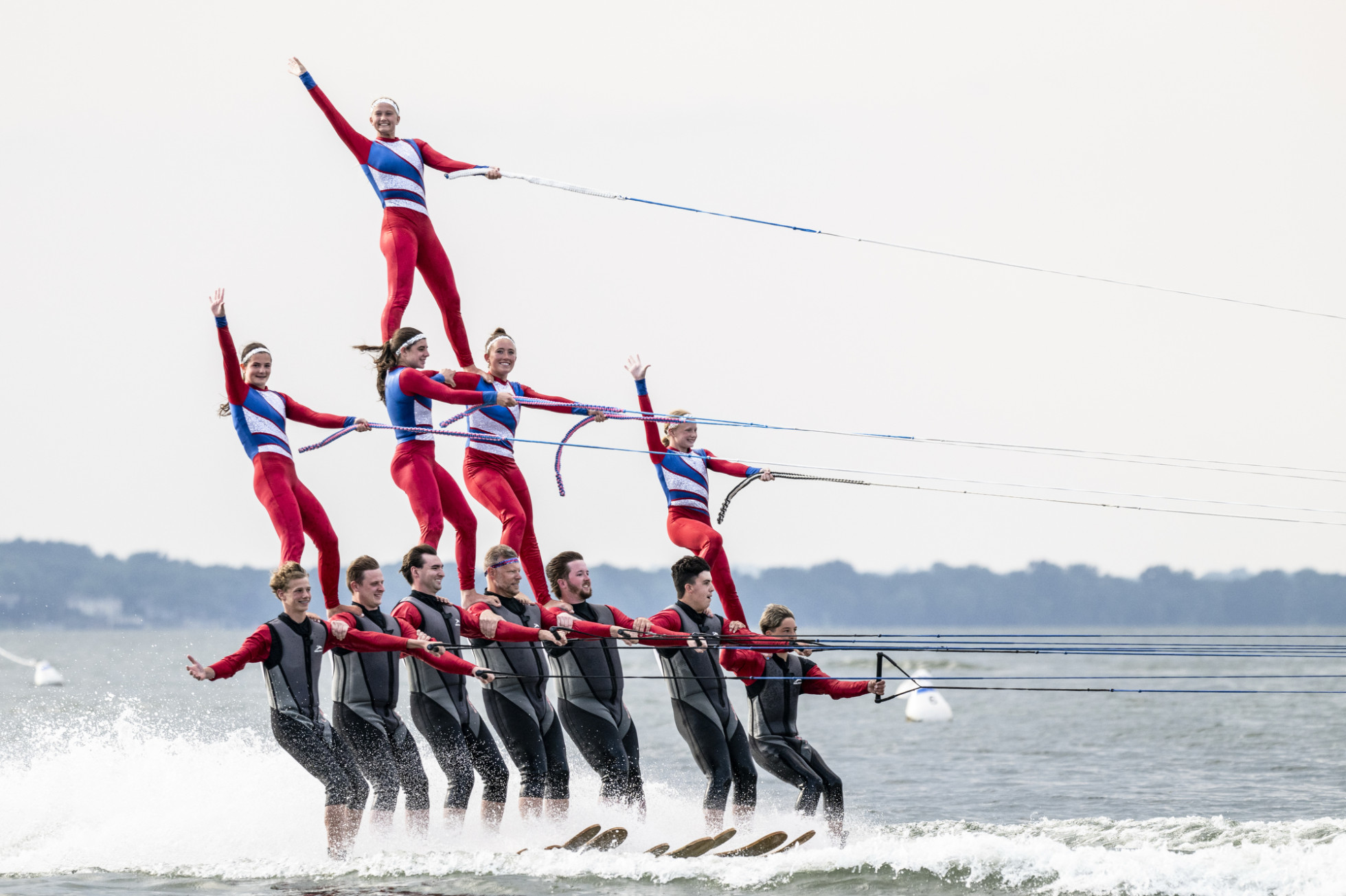 A large water ski pyramid consists of 12 performers in red, white and blue attire balancing on a lake. 