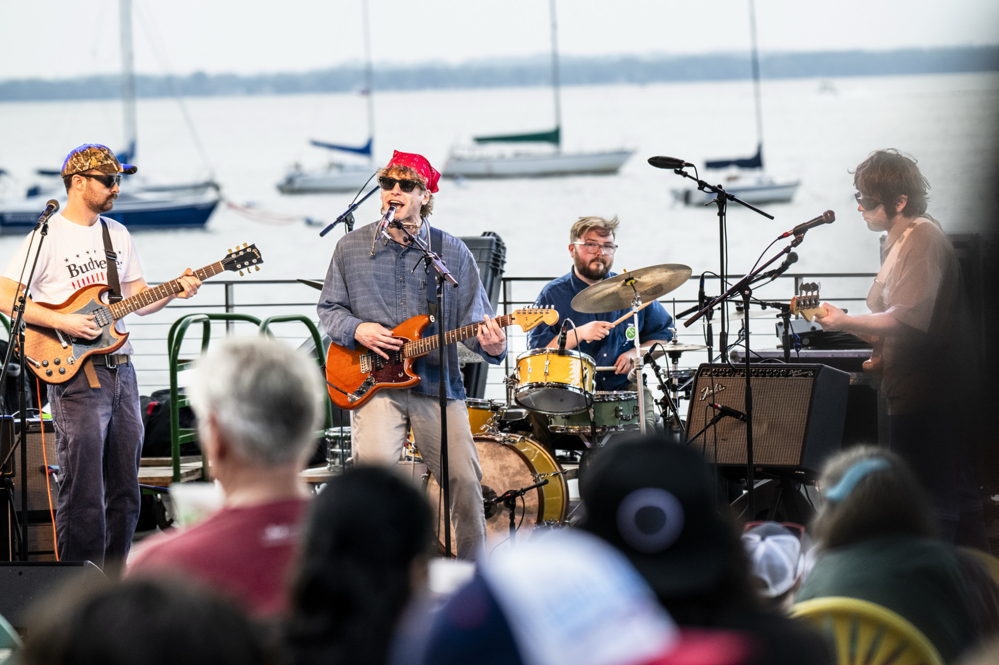A four-piece rock band performs live on an outdoor stage overlooking a lake dotted with sailboats. 
