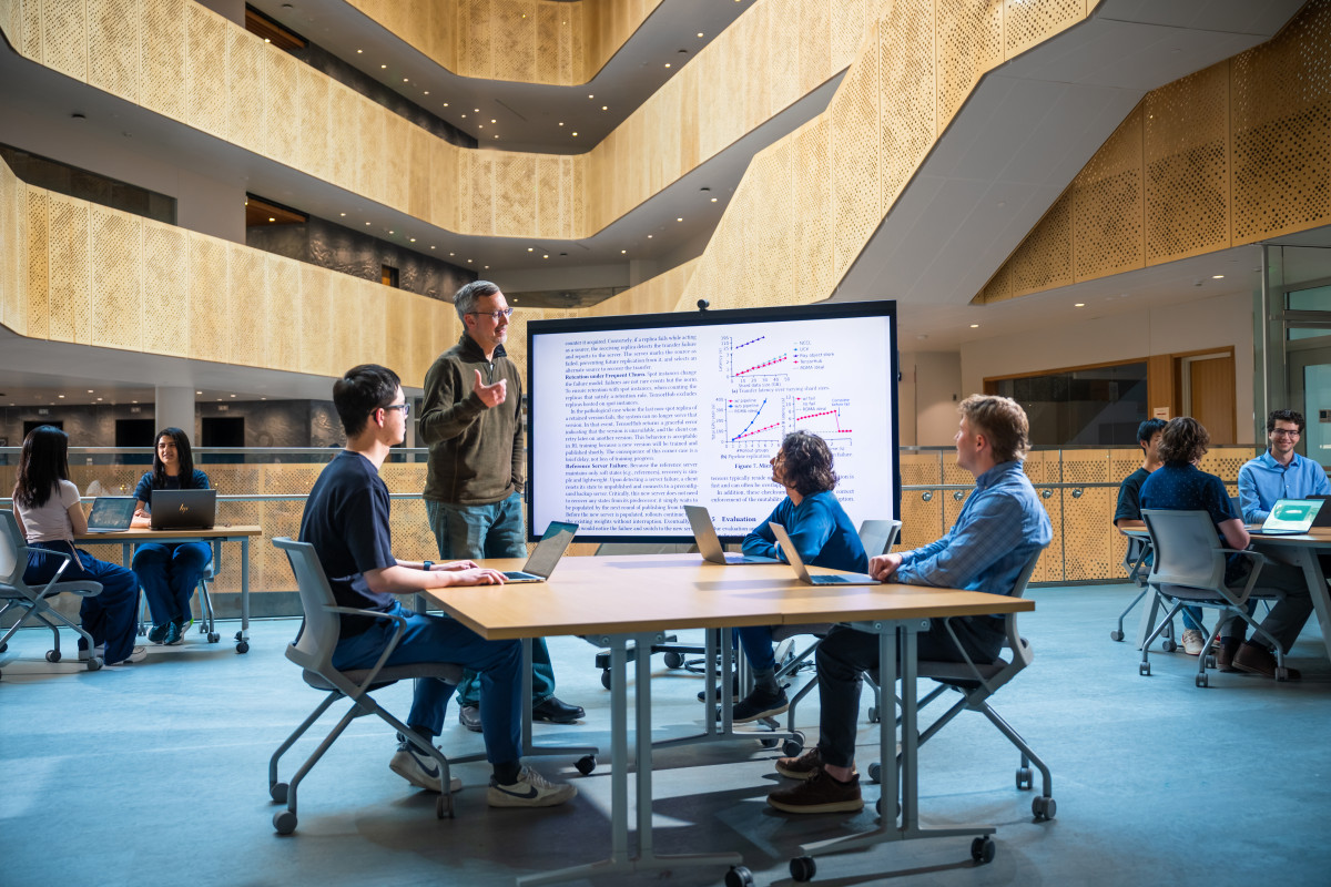 Students at tables listen to a speaker in front of a projection screen with text from a research study. 