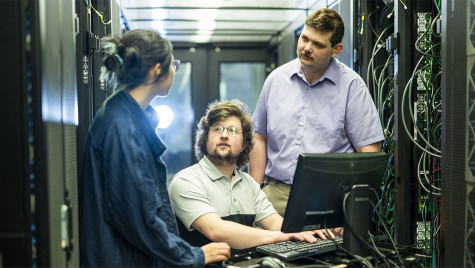 One person sits at a computer while two people stand on either side of him inside a room full of computing hardware.