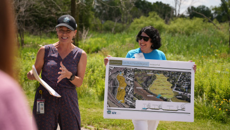 Two women stand outside talking to a group of people. One holds a large poster with a map while the other talks about the contents of the poster.