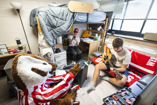 In a nearly filled residence hall room, Bucky Badger sits on the floor and listens to a student playing a guitar on his futon. The student's mother sits nearby and takes picture of the scene.