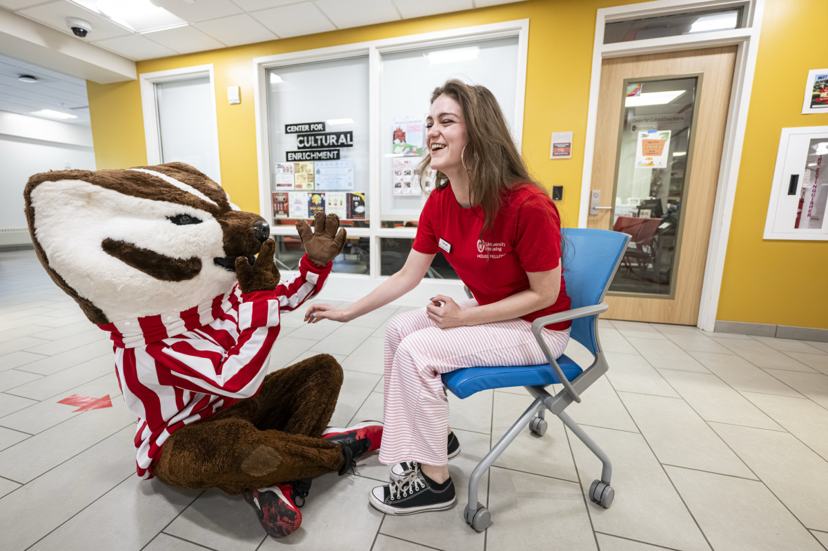 A students wears a badger red University Housing shirt and sits in a rolling chair while laughing with a playful Bucky Badger who sits on the floor in front of her.