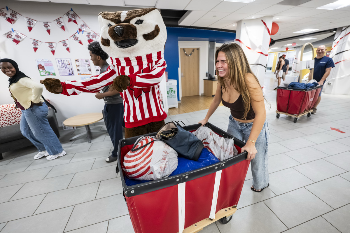 A student pushes a red moving cart filled with bags passed a playful Bucky Badger who pretends to direct traffic in the lobby of a residence hall.