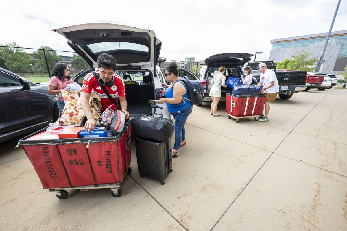 Two families stand outside of their parked cars and fill red moving carts with suitcases and furniture.