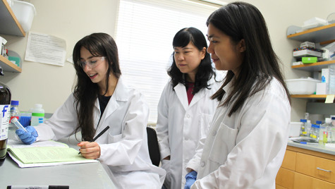 Three women wearing white lab coats stand at a counter and review the contents of a plastic vial.