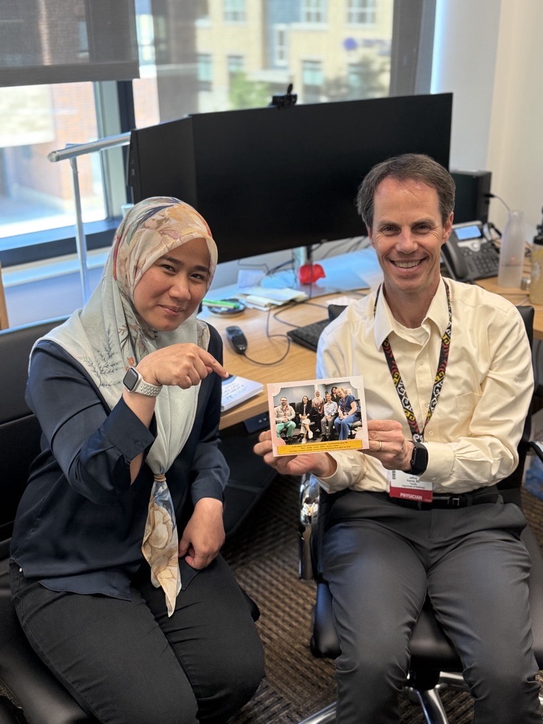 A man and a woman sit together and smile for a picture. The man is holding a photo of a group of people and the woman is pointing at the photo frame.