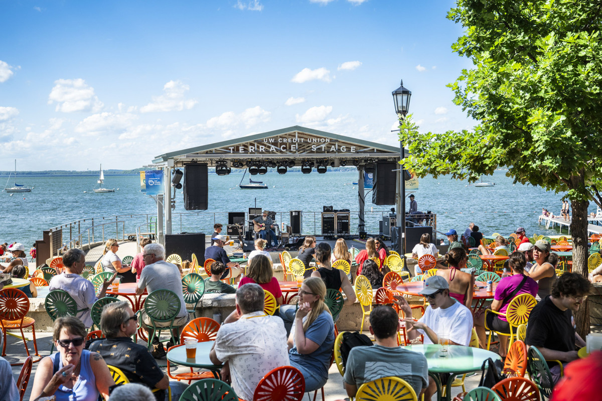 Terrace patrons listen to a performer playing guitar on stage against the Lake Mendota backdrop
