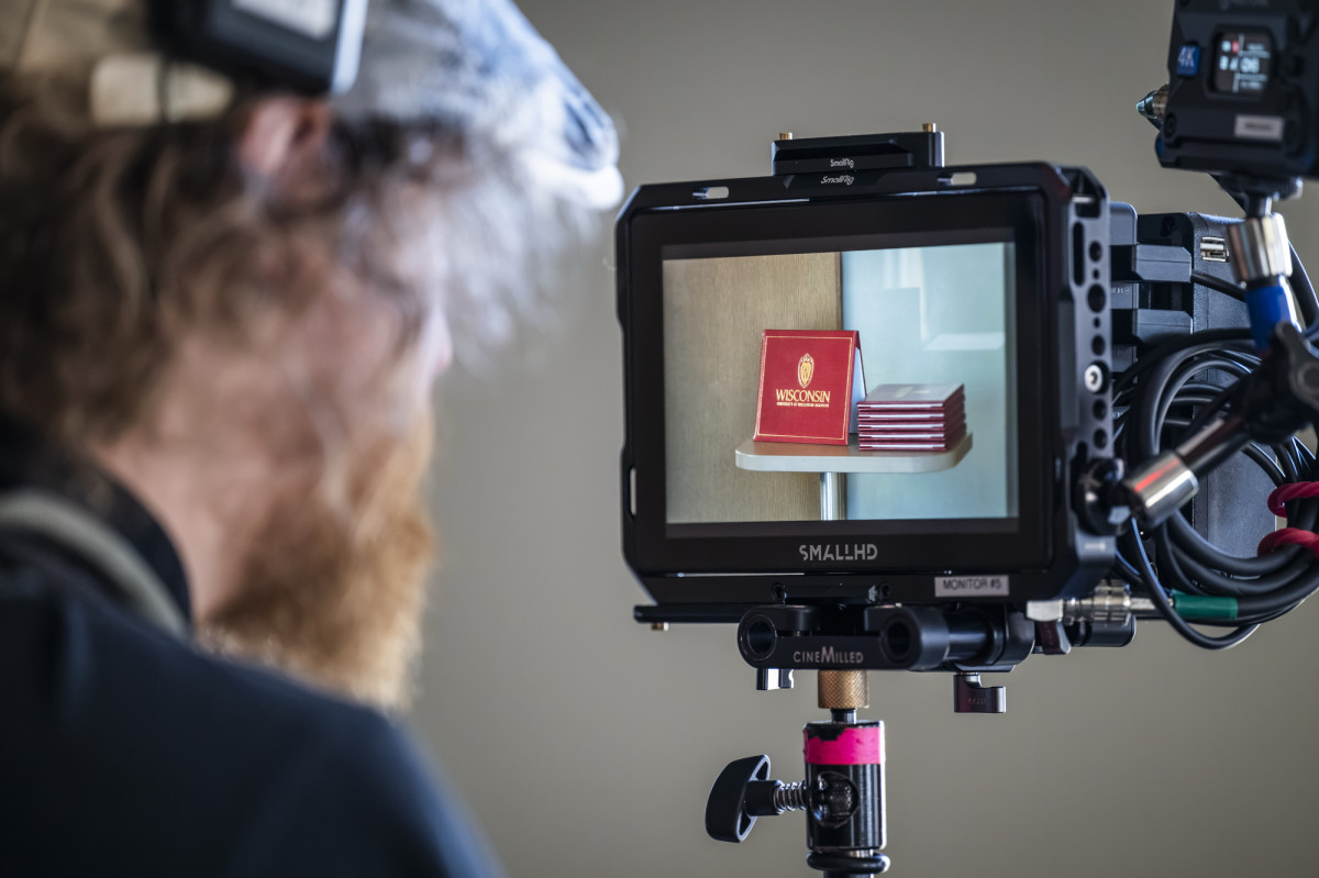 A person looking at the screen of a video camera, which is focused on a stack of red, UW–Madison diploma folders