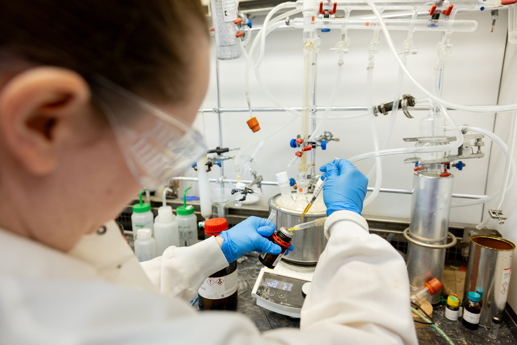 A woman wears a white lab coat, latex gloves and safety goggles while working with chemicals in a lab setting.