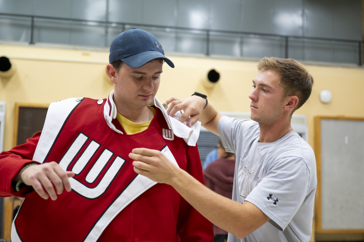 A band member helps his peer put on a marching band uniform. 