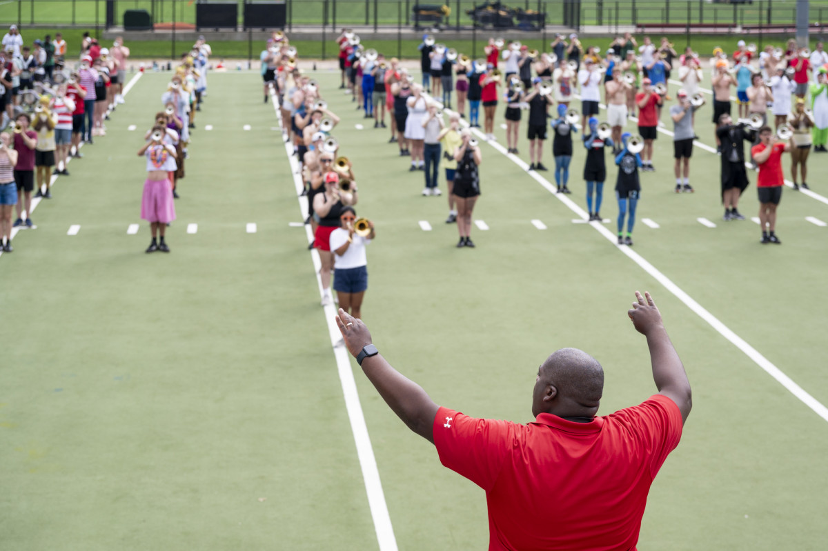 A band director conducts a large group of UW Marching Band members arranged in rows across an outdoor turf field.