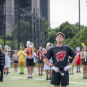 A drum major wearing a black Wisconsin shirt and white gloves stands attentively on an outdoor turf field with hands on his hips, holding a mace, with trumpet players in formation behind him.