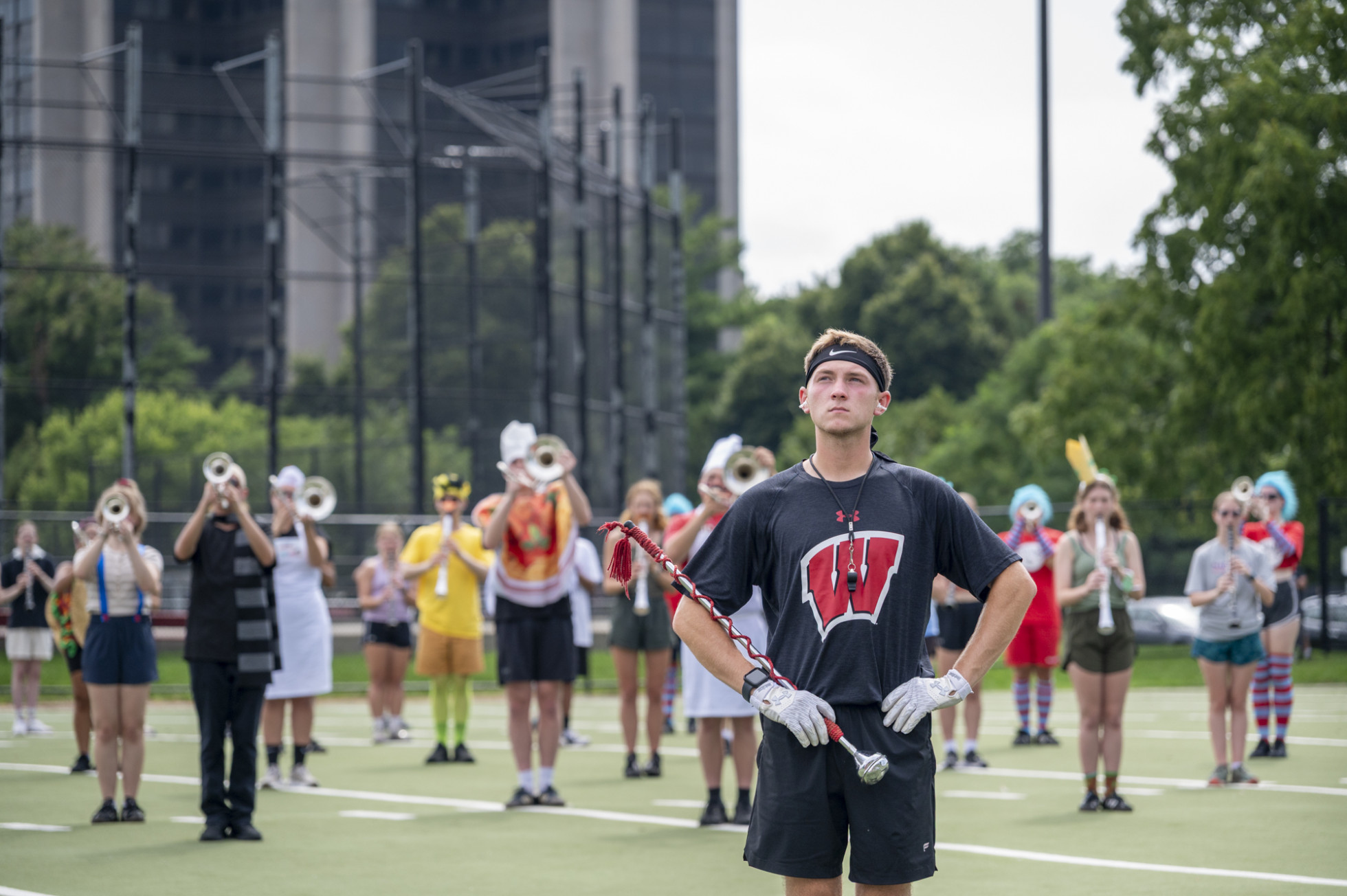 A drum major wearing a black Wisconsin shirt and white gloves stands attentively on an outdoor turf field with hands on his hips, holding a mace, with trumpet players in formation behind him.
