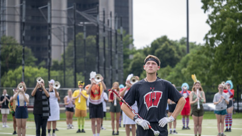 A drum major wearing a black Wisconsin shirt and white gloves stands attentively on an outdoor turf field with hands on his hips, holding a mace, with trumpet players in formation behind him.